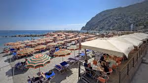 Sunny beach with rows of colorful umbrellas and sun loungers, adjacent to a cafe terrace overlooking the sea and a rocky hill.
