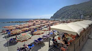 Sunny beach with rows of colorful umbrellas and sun loungers, adjacent to a cafe terrace overlooking the sea and a rocky hill.