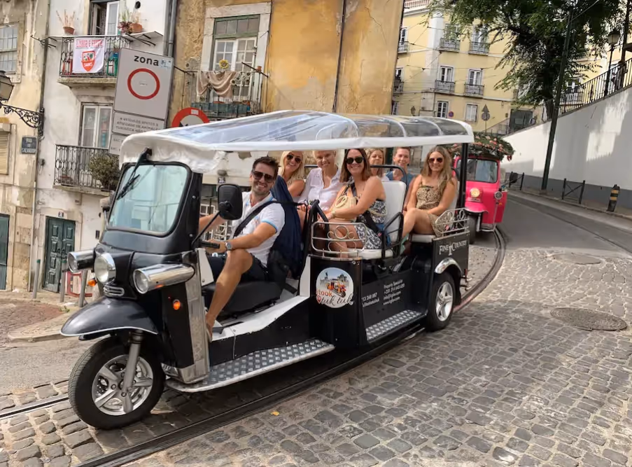 A group of seven people smiling and sitting in an open tuk-tuk vehicle on a cobblestone street in a historic urban area.