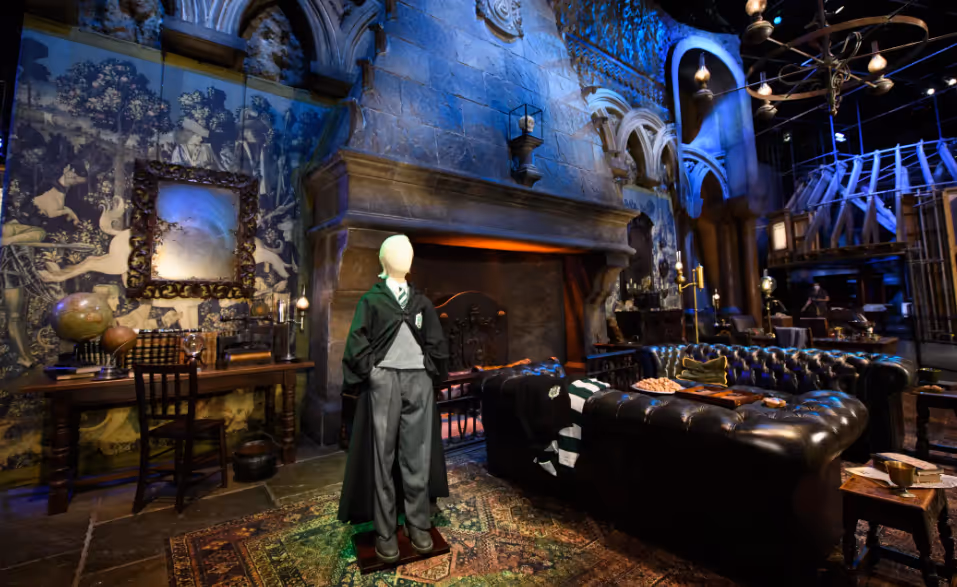 Room with stone walls, a large fireplace, a mannequin dressed in a Hogwarts-style school uniform, a patterned rug, and dark leather couches.