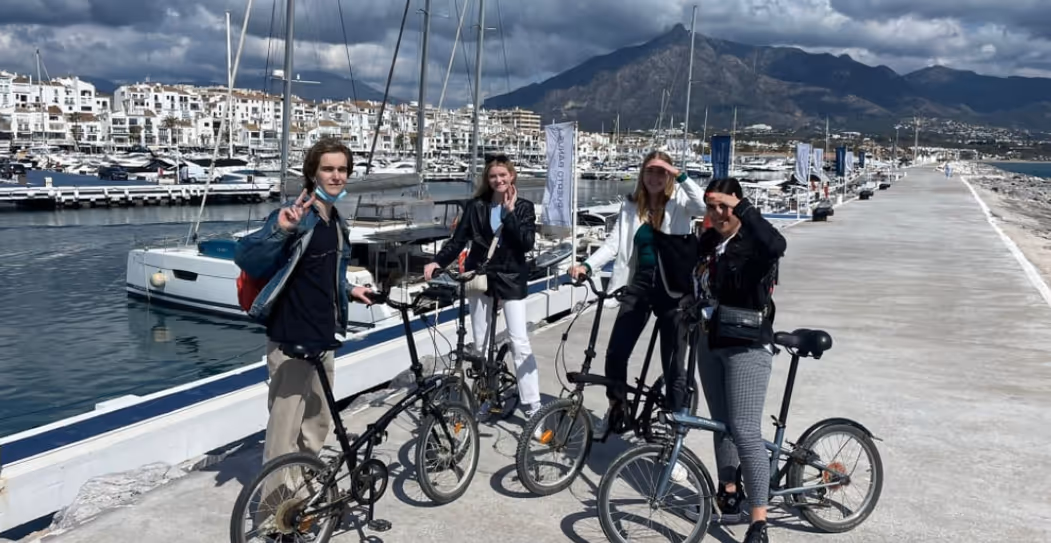 Four people posing with foldable bicycles on a marina pier with boats and mountains in the background under a cloudy sky.