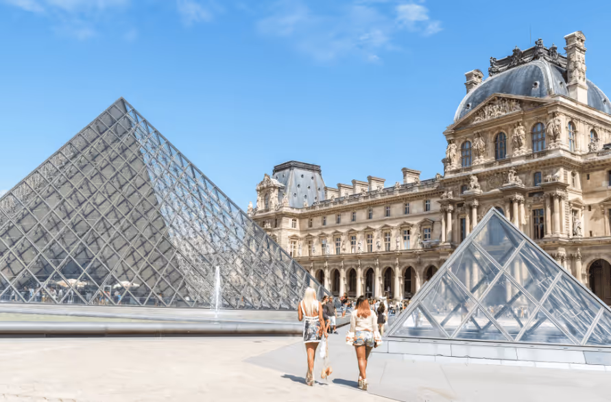 People walking near the glass pyramids of the Louvre Museum with the historic palace building in the background under a blue sky.