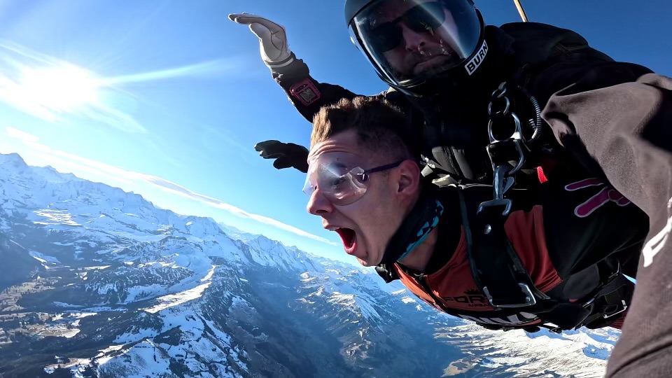 Tandem skydiving over snowy mountainous landscape with one person expressing excitement and the instructor behind wearing a helmet and goggles.