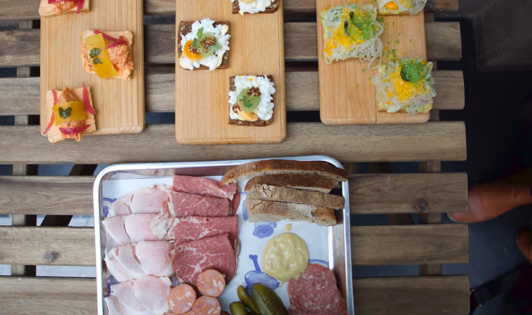 Wooden table with charcuterie tray containing various sliced meats, pickles, mustard, and bread, alongside wooden boards with assorted canapés.