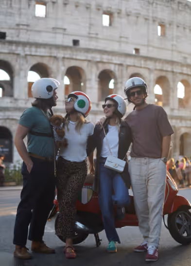Four friends wearing helmets standing in front of a red scooter with the Colosseum in Rome behind them.