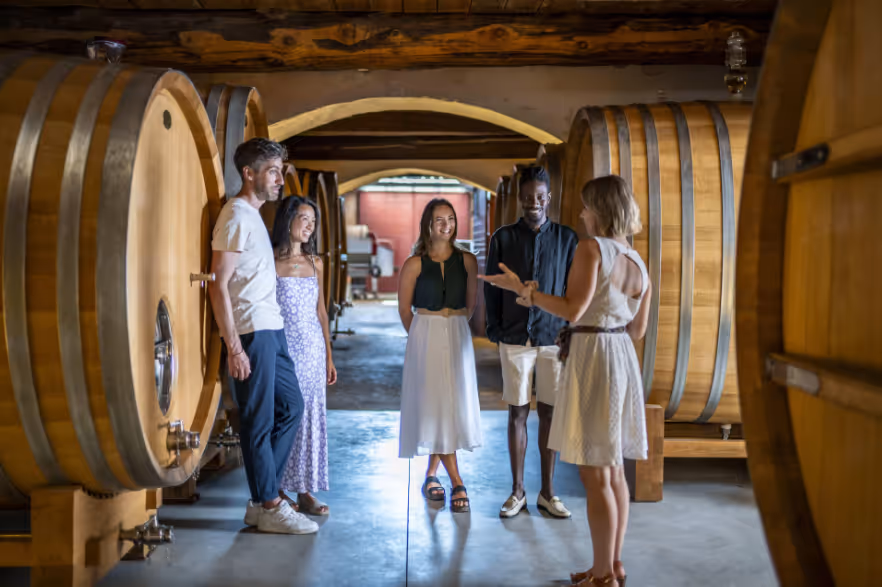A woman giving a tour to four people inside a winery cellar with large wooden wine barrels.