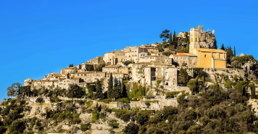 Scenic view of a hillside village with beige stone buildings and a prominent yellow church under a clear blue sky.