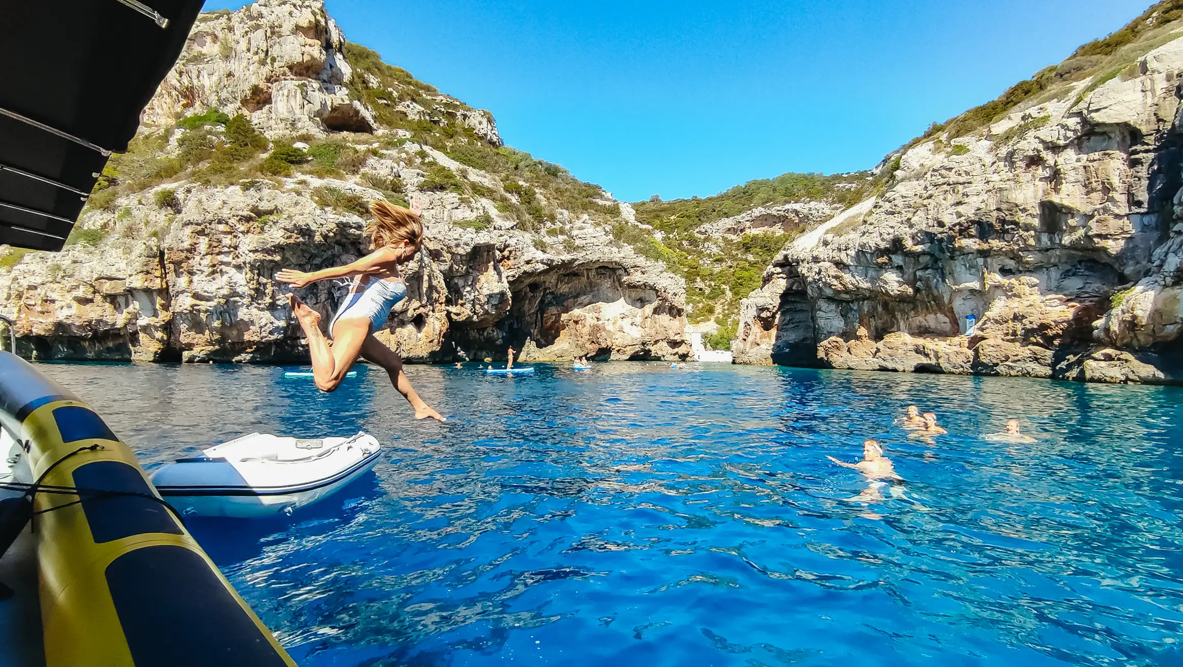 Woman in a swimsuit jumping off a boat into clear blue water surrounded by rocky cliffs with people swimming and paddleboarding.