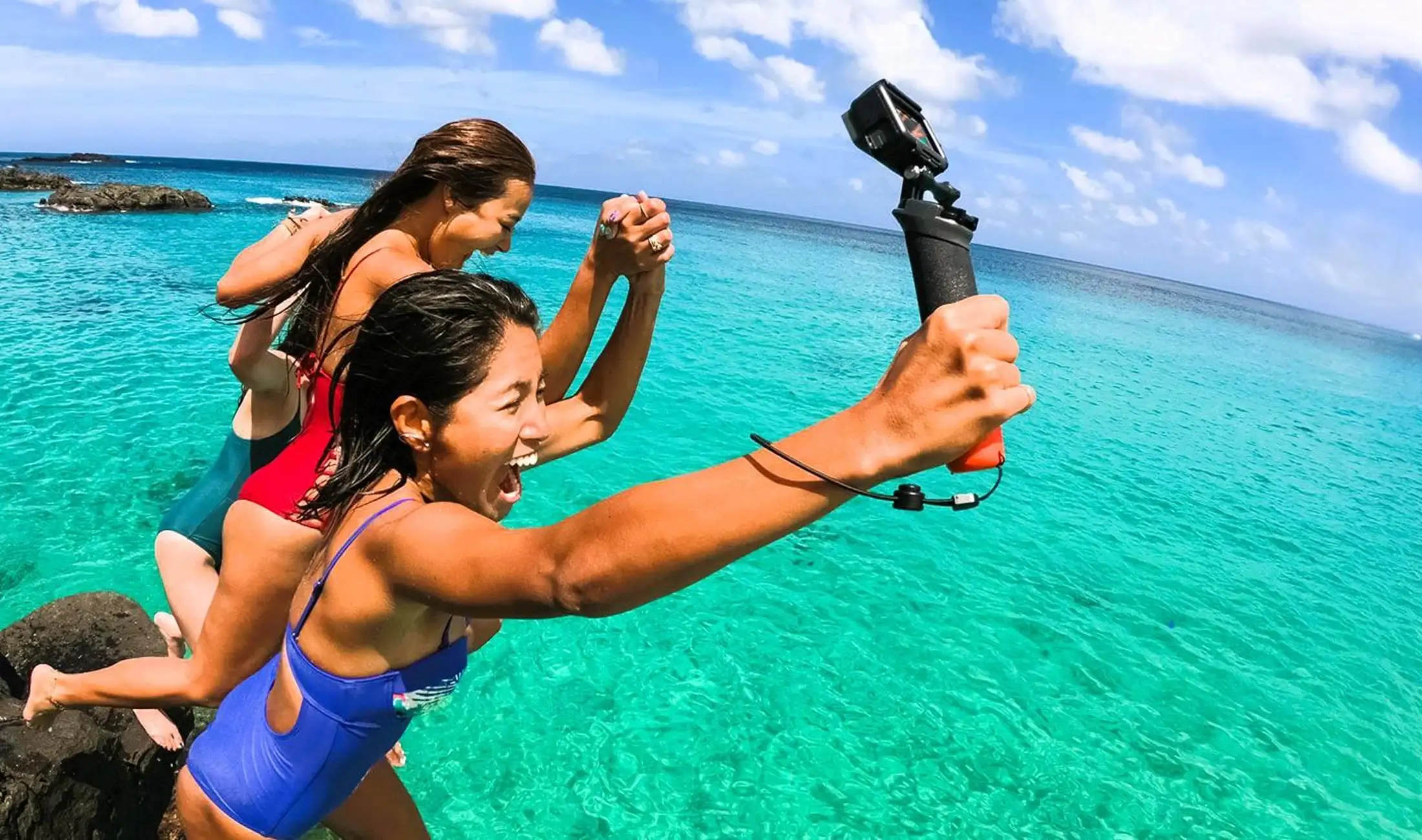 Three women holding hands and jumping joyfully into clear turquoise ocean water with a blue sky background, one holding a camera on a stick.