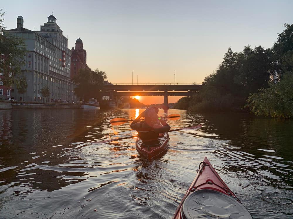 Person kayaking on a river at sunset with a bridge and buildings in the background.