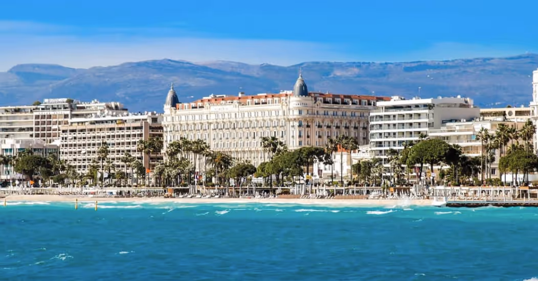 Coastal cityscape with turquoise sea, sandy beach, palm trees, and large historic buildings under a clear blue sky.