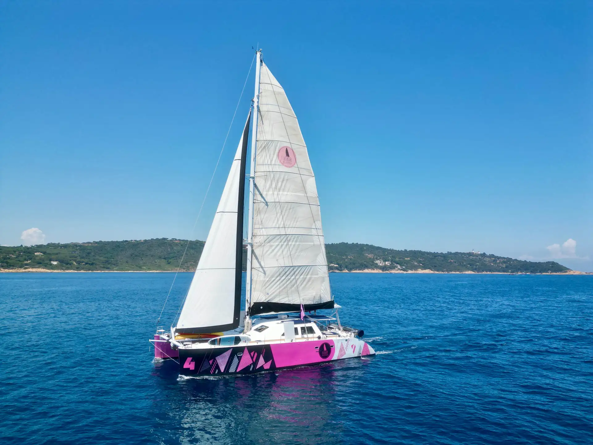 Pink and white catamaran sailing on calm blue sea with a clear sky and green coastline in the background.
