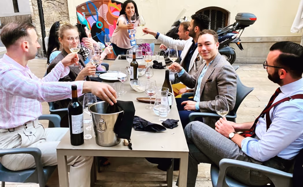 Group of seven people sitting around a table outdoors enjoying a wine tasting and toasting with wine glasses.