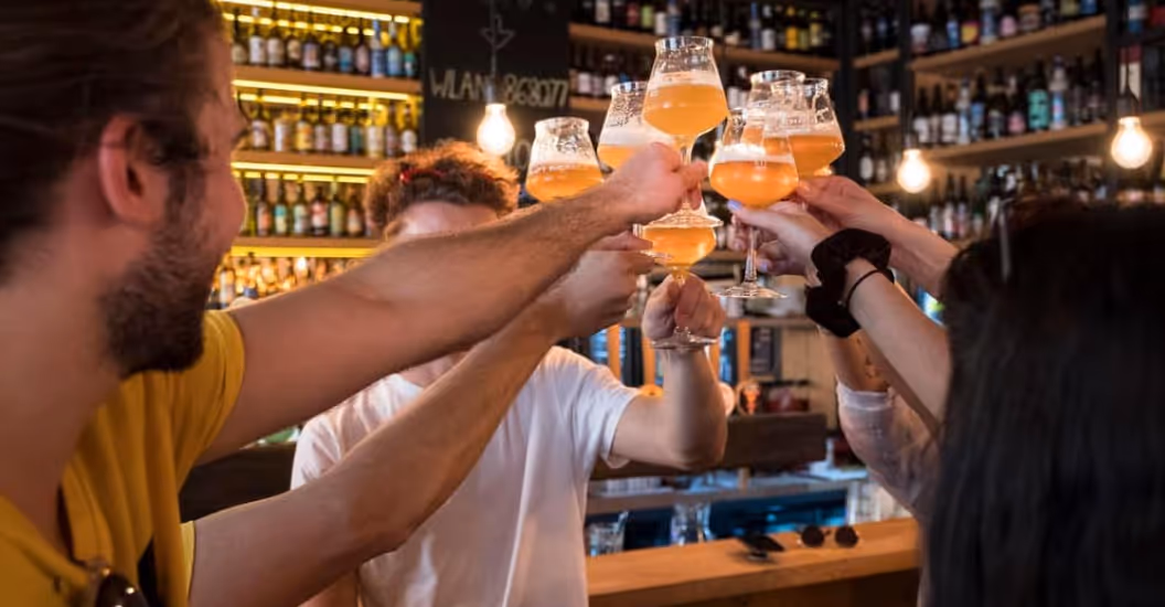 Group of friends clinking glasses of craft beer in a bar with shelves of beer bottles in the background.