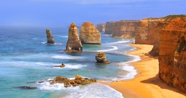 Coastal view of the Twelve Apostles limestone stacks along a sandy beach and blue ocean under a clear sky.