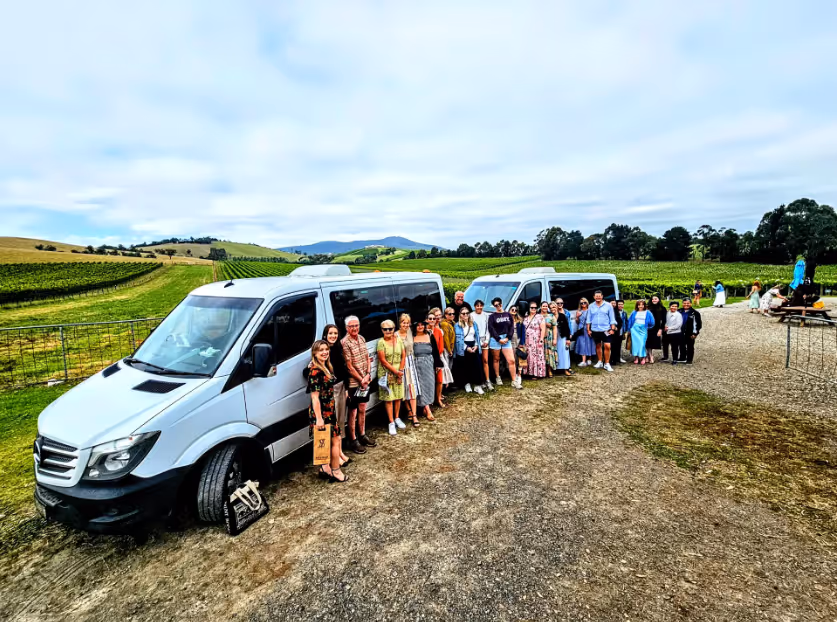 Group of people standing in a line beside two white vans on a gravel path with green fields and hills in the background under a cloudy sky.