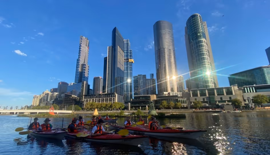 Group of kayakers wearing life jackets paddling on a river with a city skyline and tall modern buildings in the background under a clear blue sky.