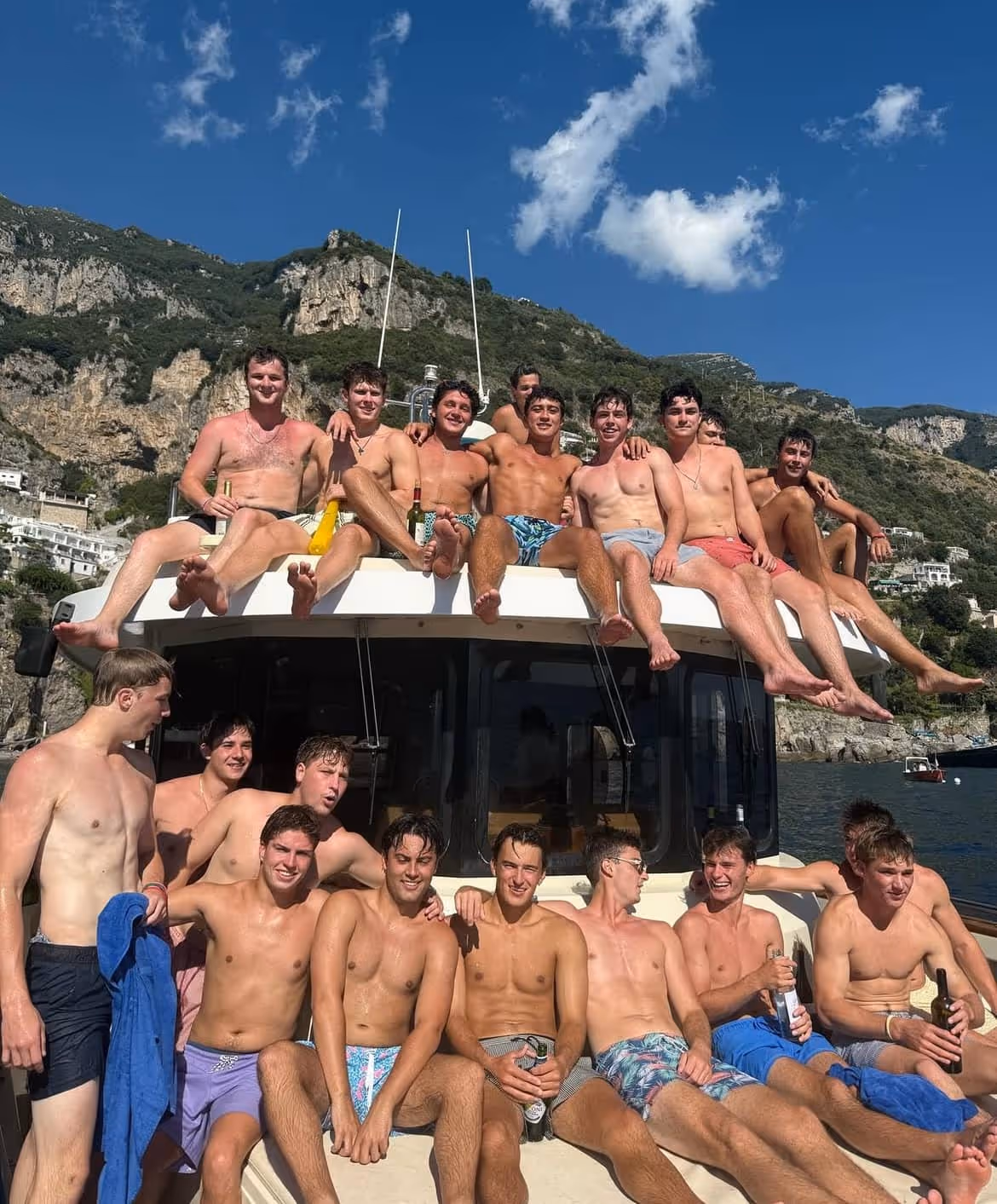 Group of young men in swim trunks sitting and smiling on a boat with rocky hills and a blue sky in the background.