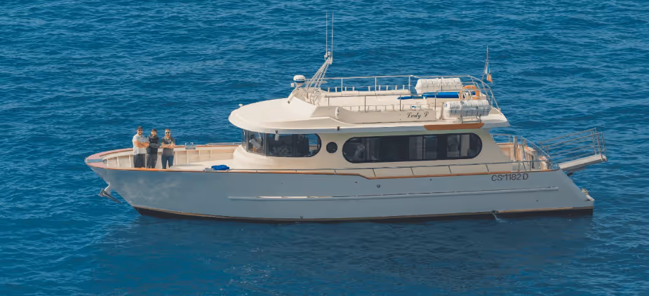 White motor yacht named Lady V floating on calm blue ocean water with three people standing at the bow.