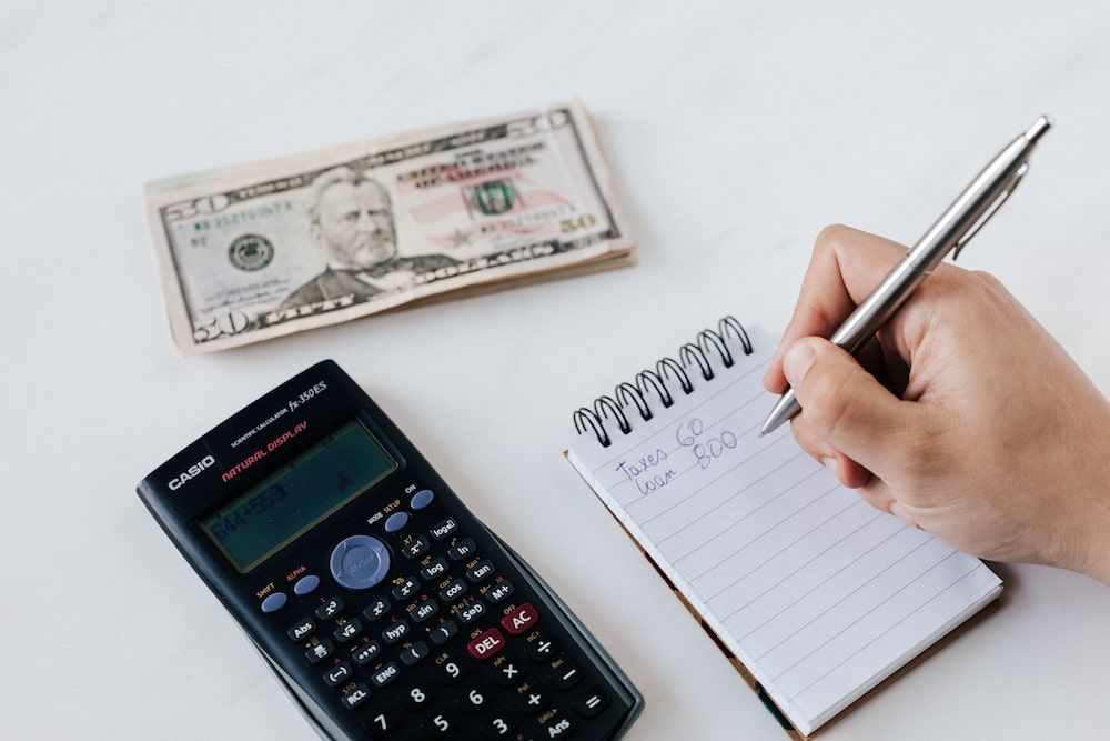 Calculator, stack of money, and notepad with someone taking notes 