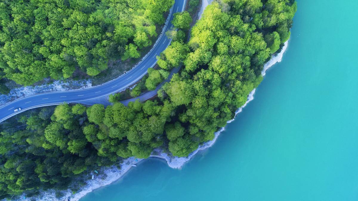 A curved road running through dense green forest next to a turquoise water body.