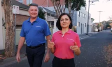 A man in a blue shirt and a woman in a pink shirt walking outdoors on a tree-lined street with buildings in the background, both smiling and giving thumbs up.