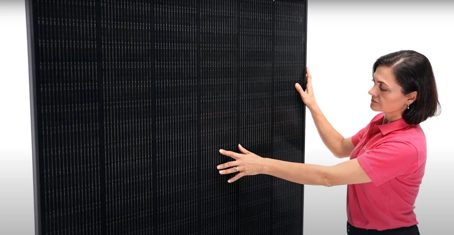 Woman in a pink shirt examining a large black solar panel, touching its surface with both hands.