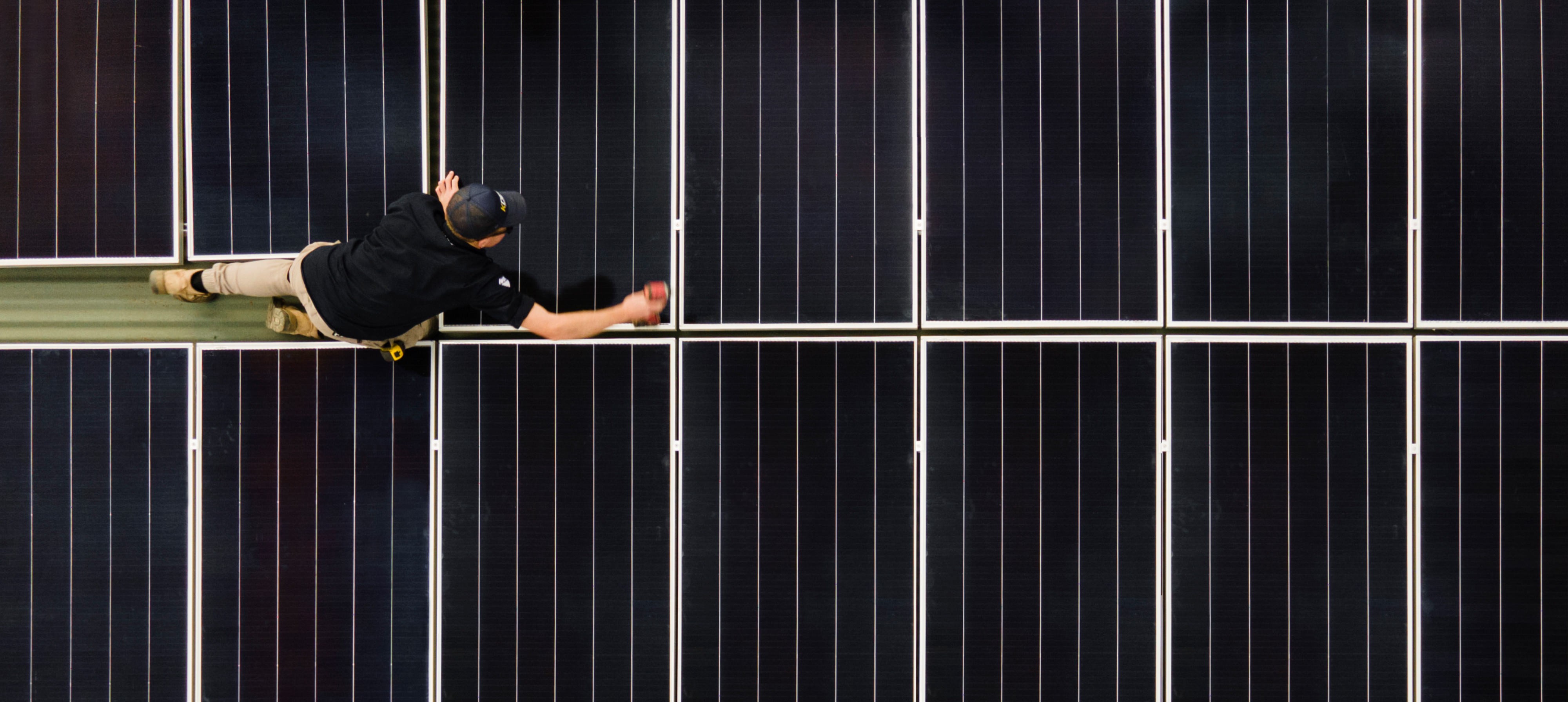 A man in a black shirt and beige pants installing or inspecting black solar panels from above, reaching out with his right hand.
