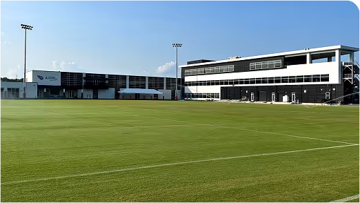 Modern athletic training facility with well-maintained grass field under a clear blue sky, equipped with stadium lighting — ideal for professional sports teams and field management.