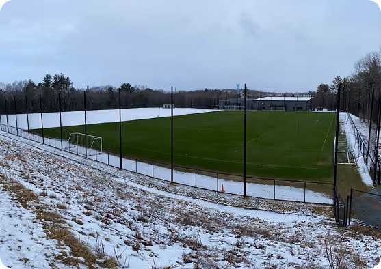 Snow-lined view of the New England Revolution soccer training grounds with fenced fields and surrounding forest in winter.