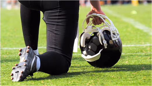 Football player kneeling on grass field holding helmet during pregame warm-up, close-up of gear and cleats