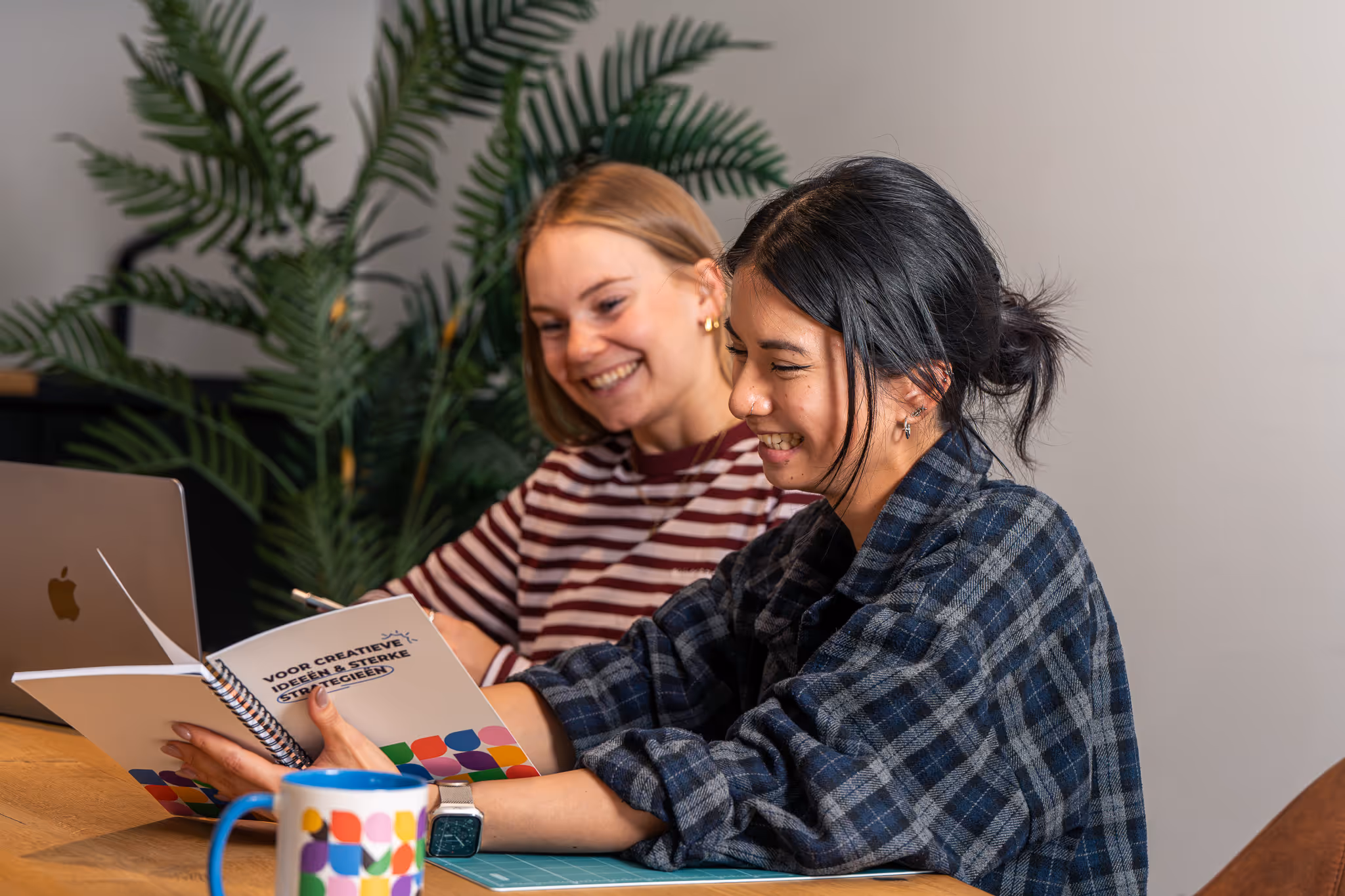 Twee vrouwen lezen samen een notitieboek met de titel 'Voor creatieve ideeën & sterke strategieën' terwijl ze aan een tafel zitten met een laptop en een kleurrijke mok.