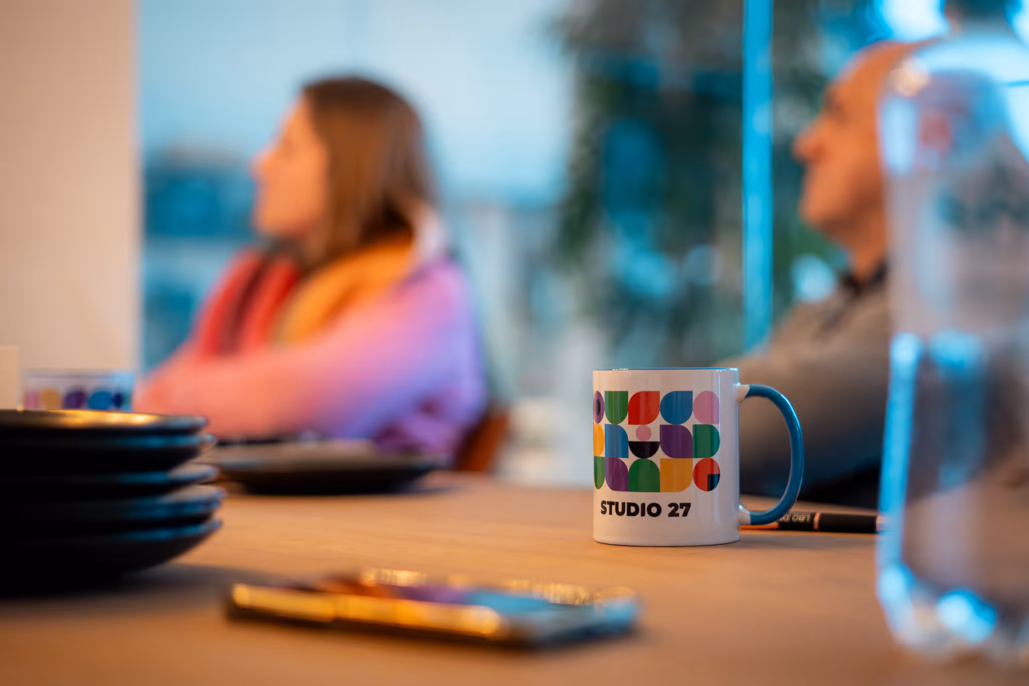Een mok met een kleurrijk patroon en de tekst Studio 27 op een tafel, met wazige mensen op de achtergrond.
