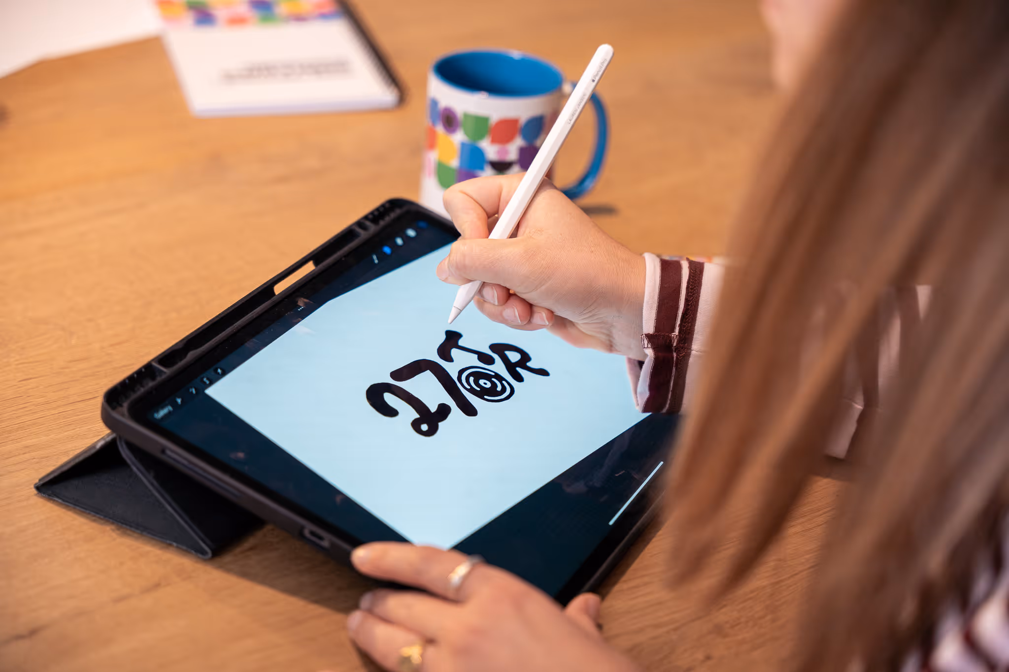 Persoon tekent met een digitale pen een zwart ontwerp op een tablet met een houten tafel en een kleurrijke mok op de achtergrond.