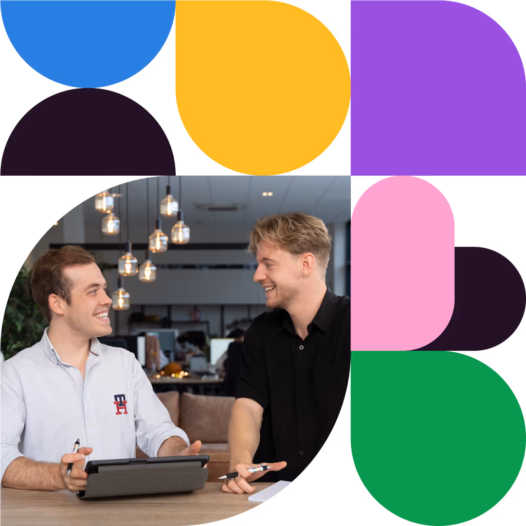 Twee jonge mannen zitten aan een tafel in een kantoor en lachen terwijl ze met een tablet en pennen werken.