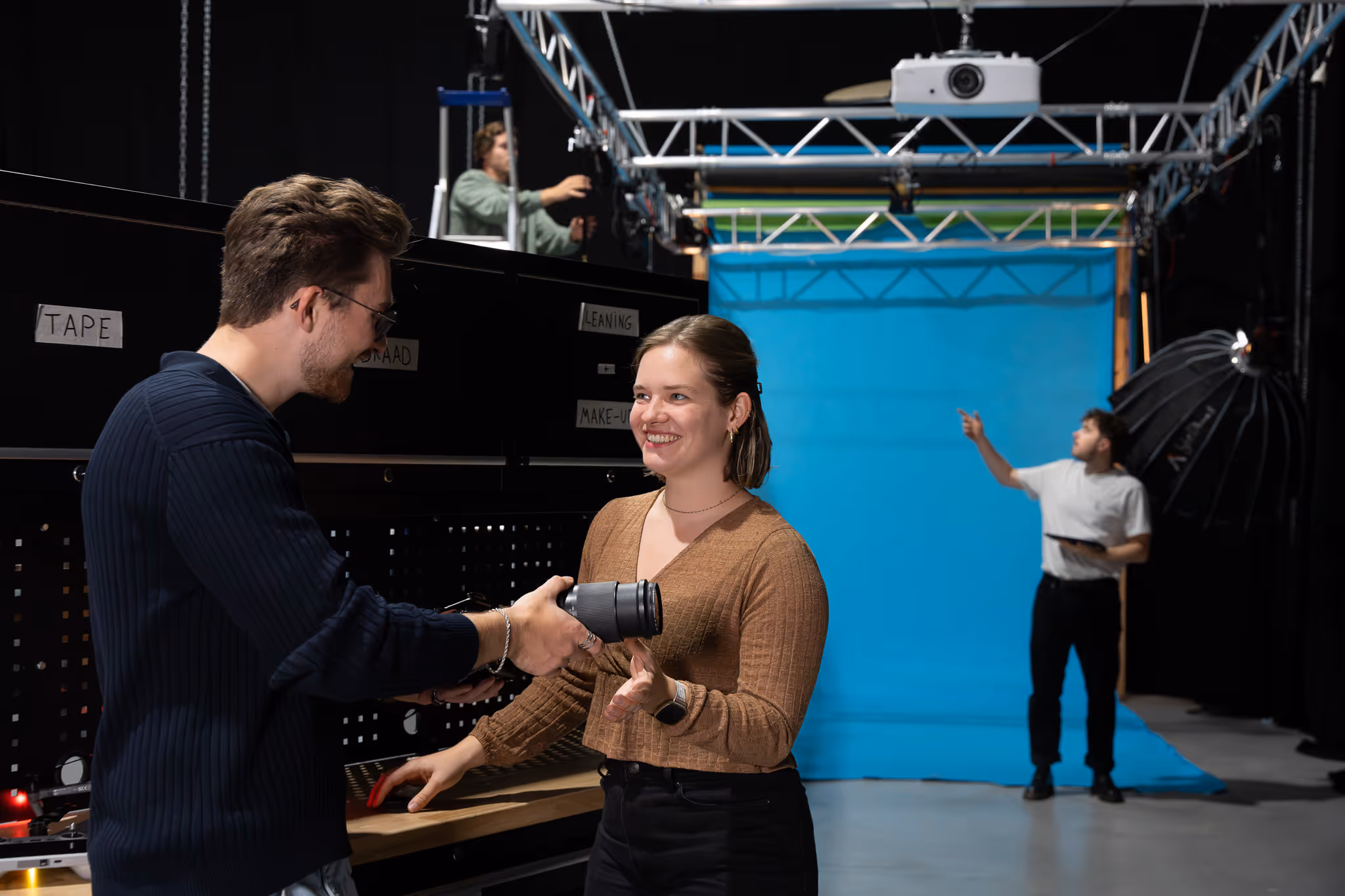 Twee mensen wisselen een cameralenzen uit in een fotostudio met een blauwe achtergrond en apparatuur.