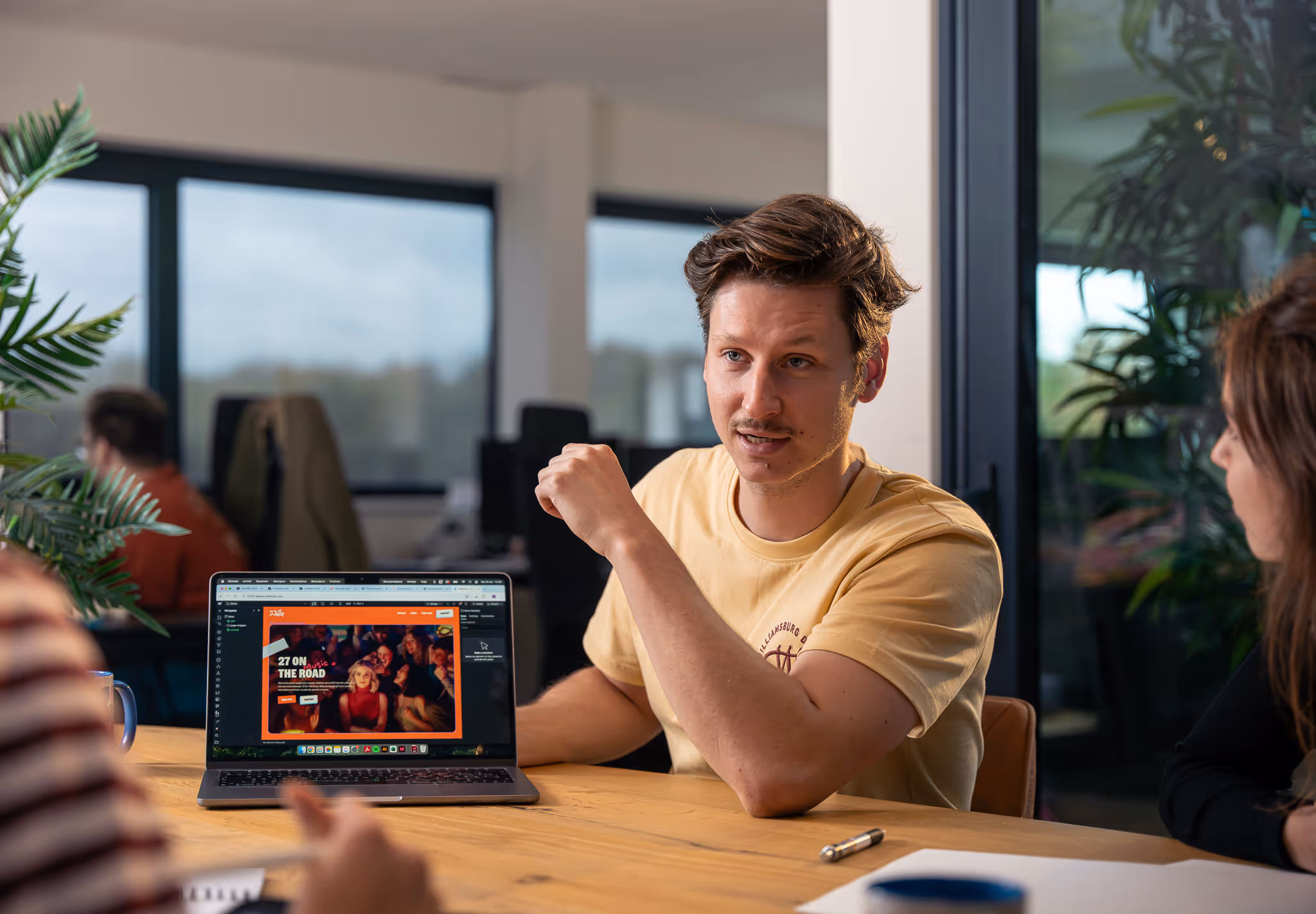 Man in geel T-shirt die aan een tafel zit en een gesprek voert, met een open laptop voor zich waarop een website zichtbaar is.