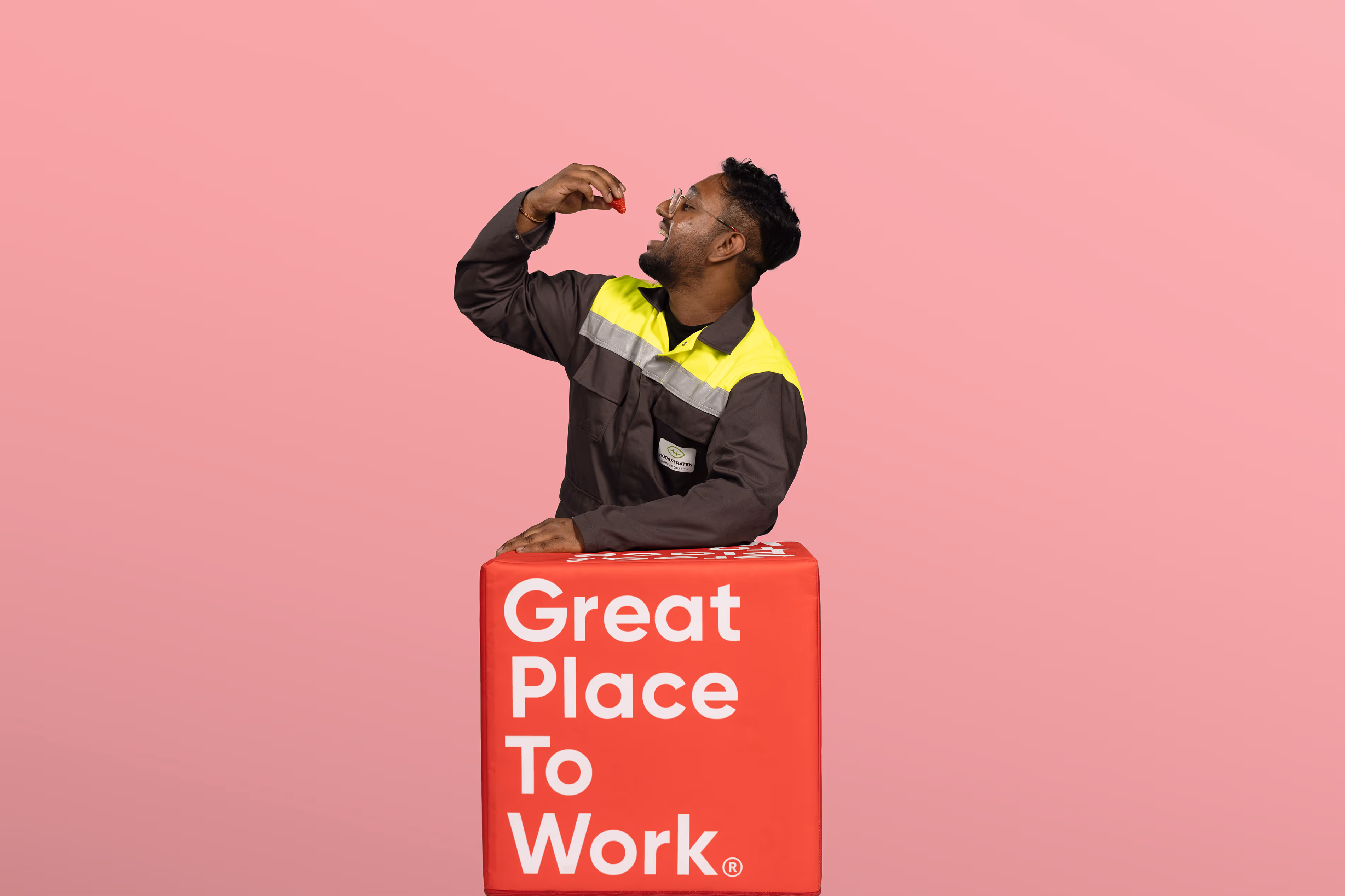 Man in werkuniform houdt een aardbei boven zijn mond met een rood blok met tekst Great Place To Work.