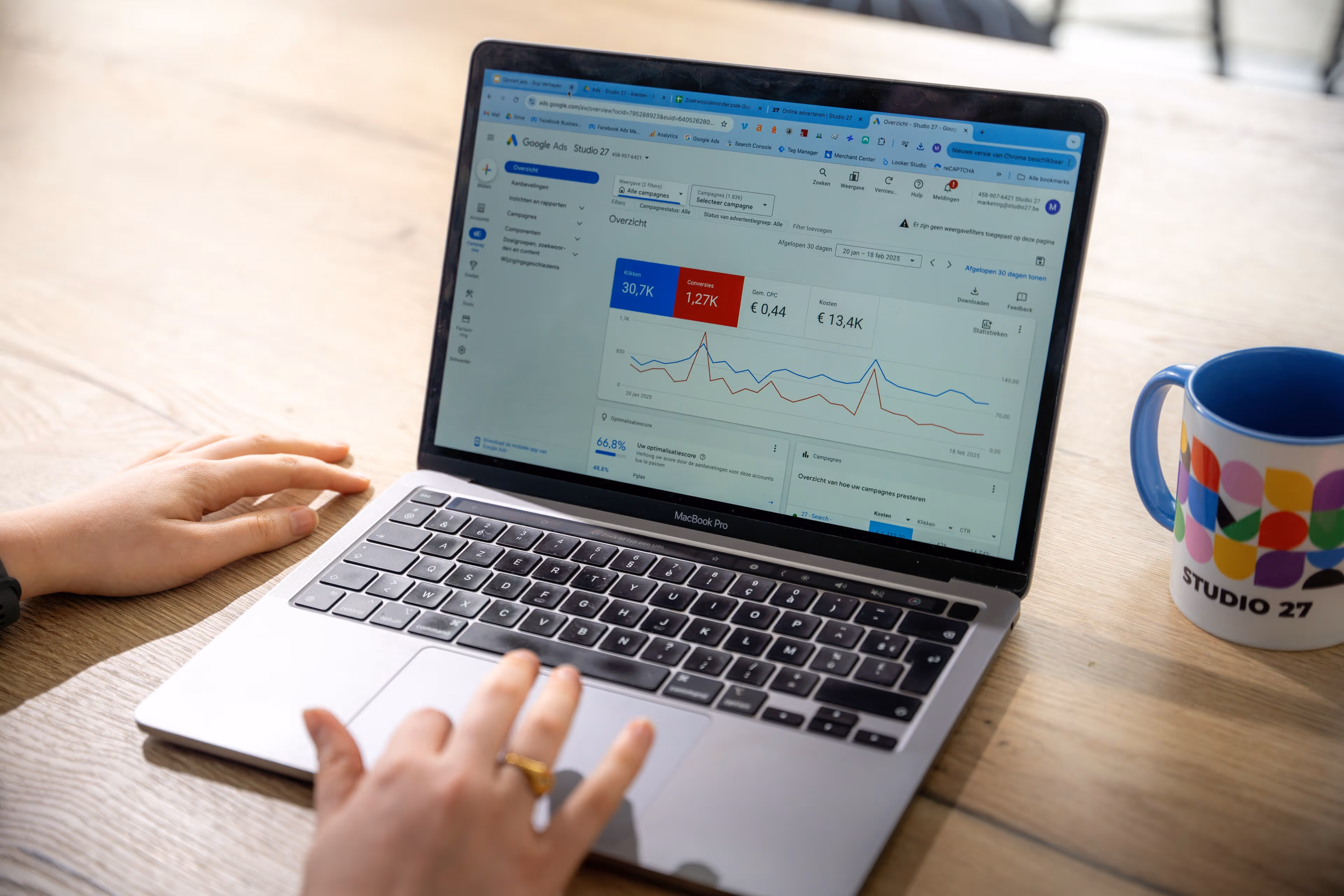 Iemand kijkt naar een laptop met een Google Ads dashboard op het scherm en een mok met Studio 27-logo op een houten tafel.