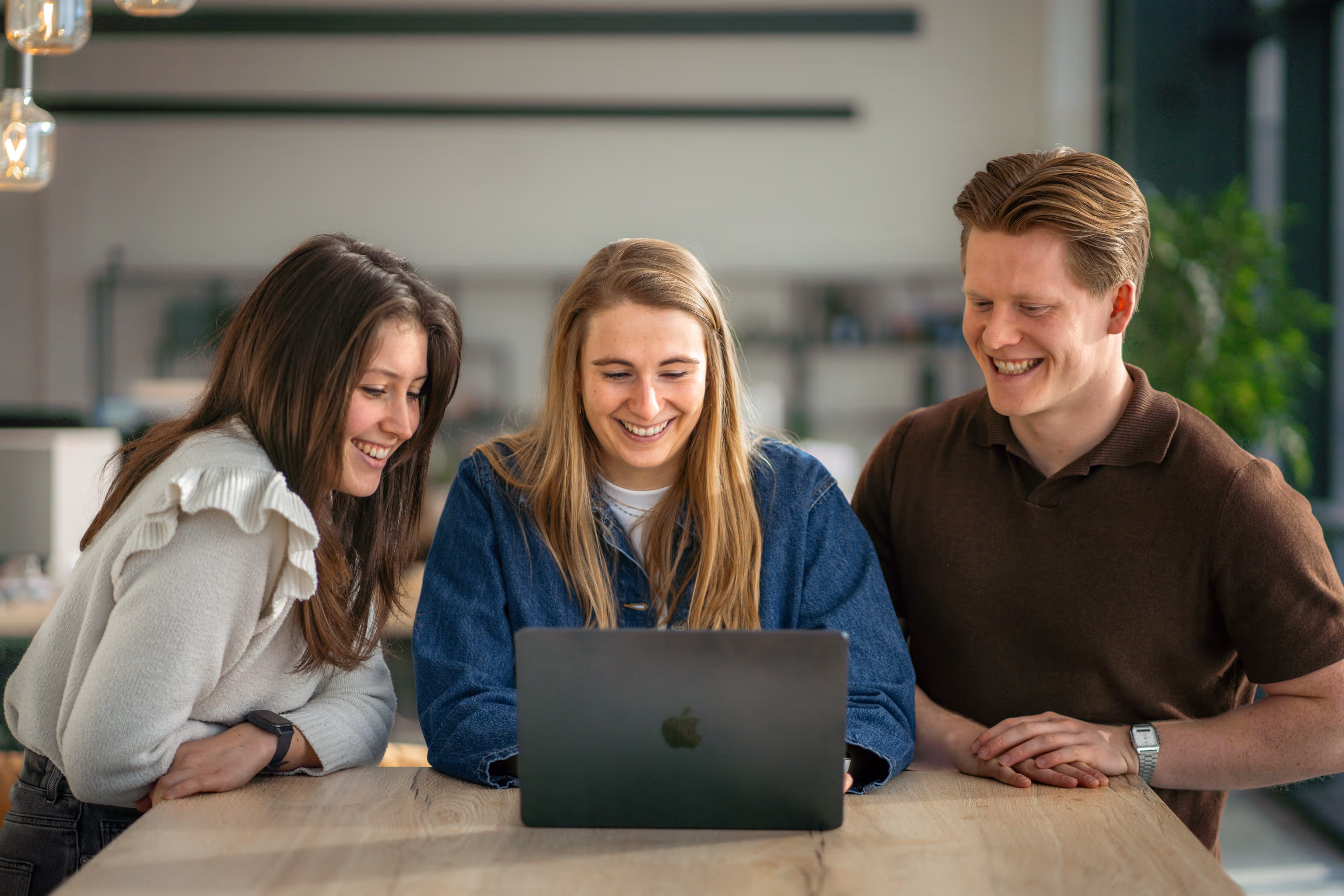 Drie jonge volwassenen lachen terwijl ze samen naar een laptop kijken aan een houten tafel.