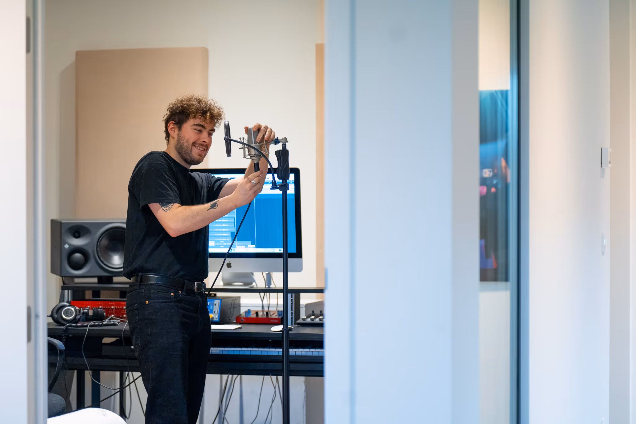 Man met krullend haar stelt een microfoon in in een opnamestudio met computer en audioapparatuur.