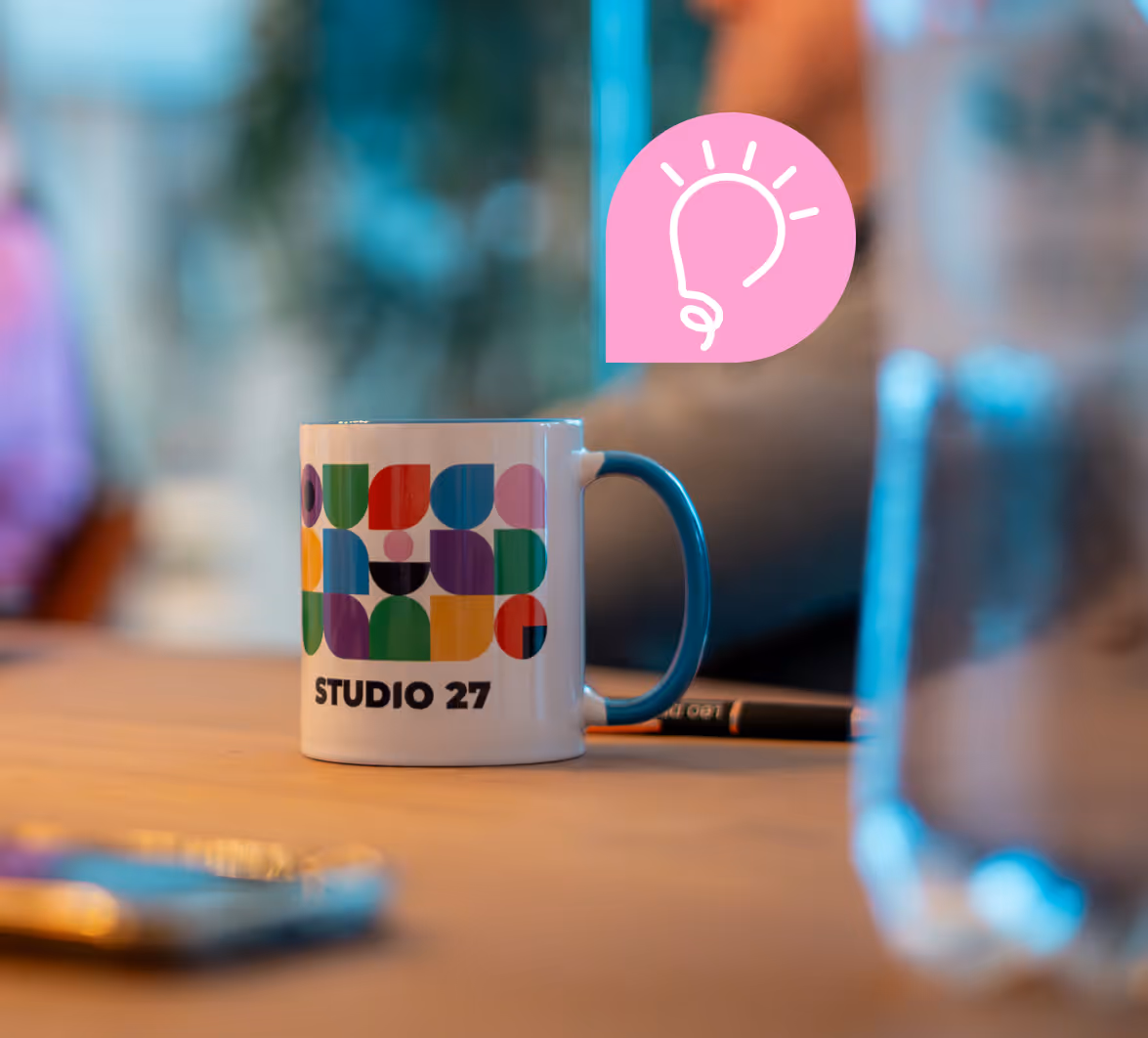 Witte koffiemok met blauwe handgreep en kleurrijk abstract ontwerp met de tekst 'STUDIO 27' op een tafel met vage achtergrond.