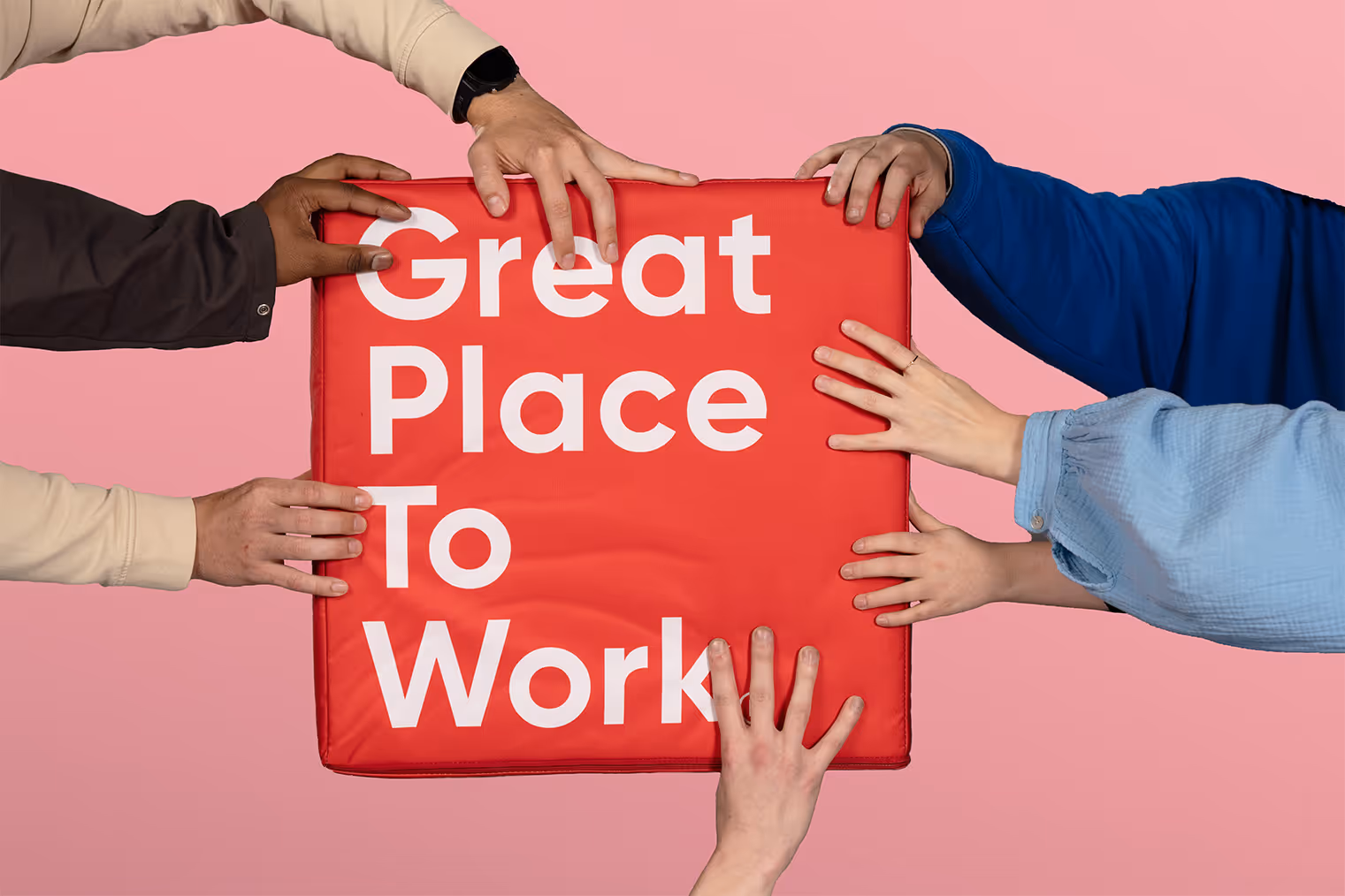 Verschillende handen houden een rood bord vast met de tekst 'Great Place To Work' op een roze achtergrond.