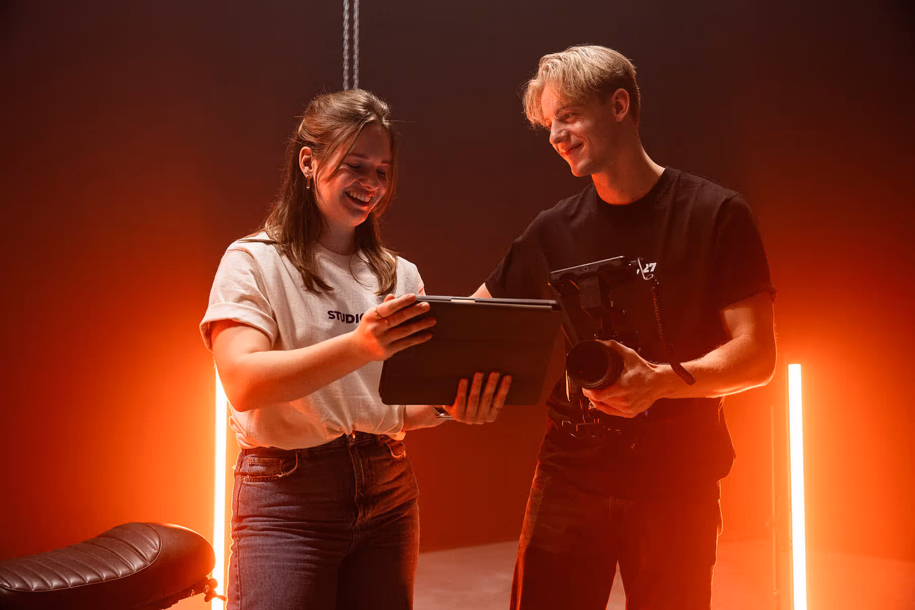 Twee jonge mensen in een studio met oranje achtergrondlicht bekijken samen een tablet terwijl een persoon een camera vasthoudt.