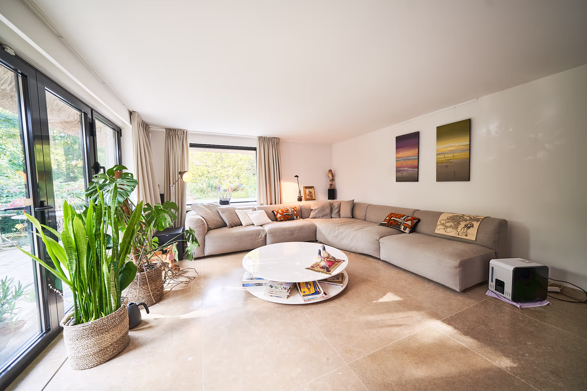 Ruime lichte woonkamer met grote beige hoekbank, ronde witte salontafel, groene kamerplanten en grote ramen met uitzicht op tuin.
