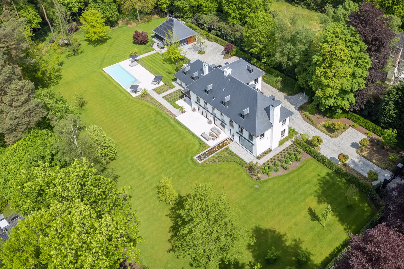 Luchtfoto van een groot wit landhuis met donker dak, omgeven door een ruim onderhouden gazon, een zwembad, een terras met tuinmeubilair en veel groene bomen.