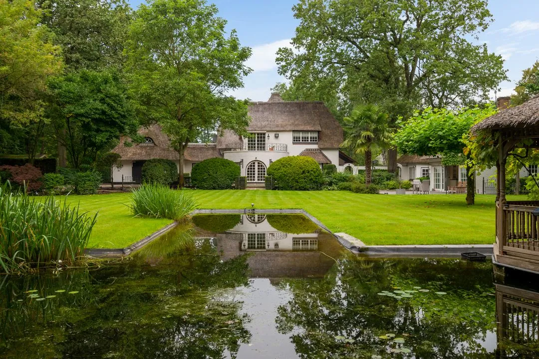 Grote tuin met een vijver en weerspiegeling van een huis met rieten dak, omringd door groene bomen en struiken.