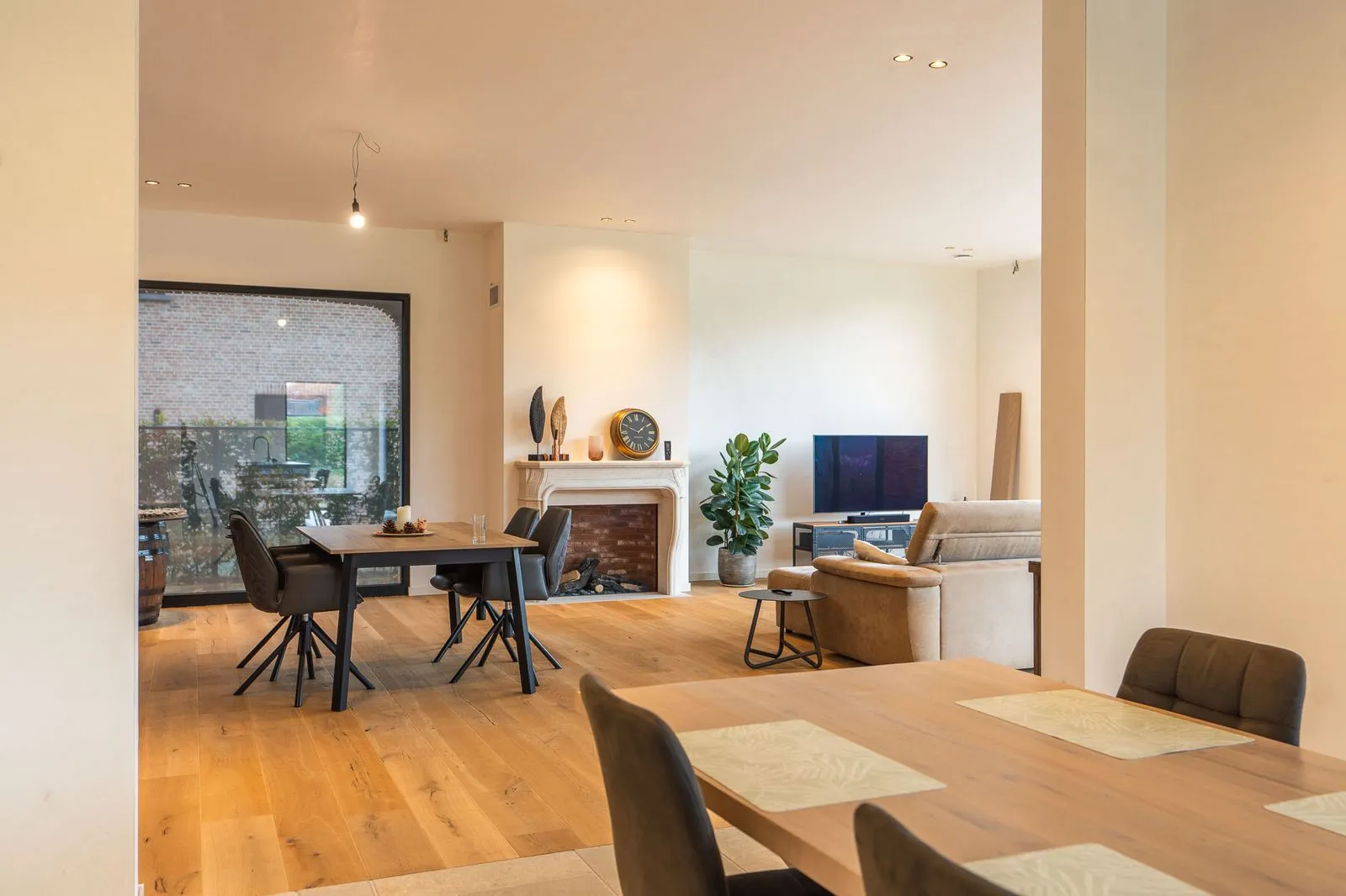 Ruime woonkamer met houten vloer, een eettafel met stoelen, een open haard met een klok, een groene plant en een beige bank met tv.