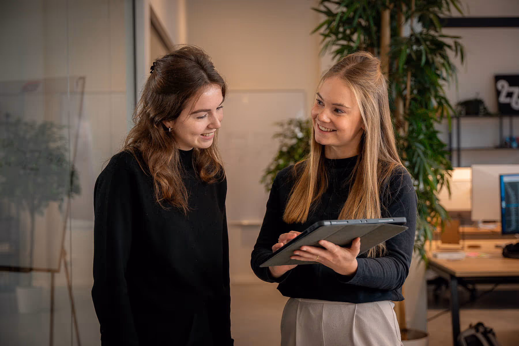 Twee jonge vrouwen in een kantooromgeving die glimlachen en naar een tablet kijken.