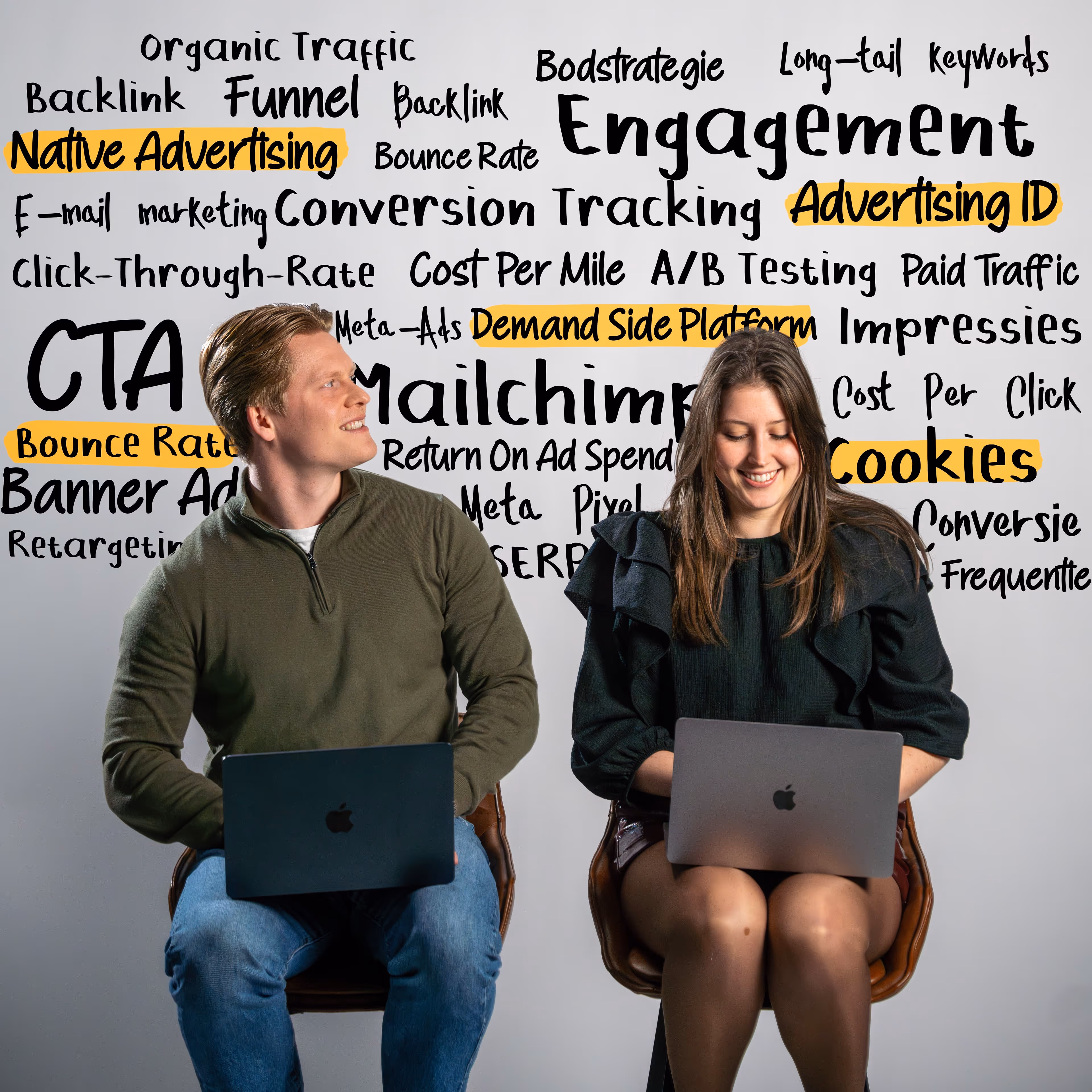 Man and woman sitting with laptops in front of a white wall covered with marketing and advertising terms, some highlighted in orange.